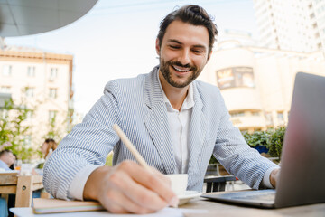 Young man wearing jacket writing down notes while working with laptop