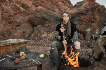 Young homeless woman sitting in front of the fire and cooking the meal for herself outdoors
