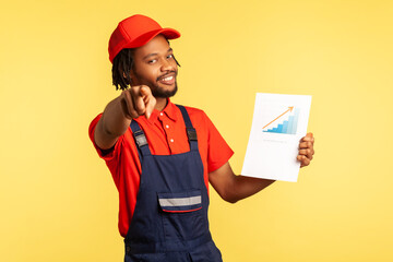 Positive handyman in overalls holding paper chart showing increase of workers salaries, pointing to...