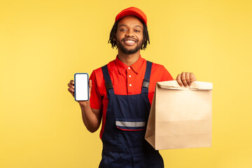 Delivery service mobile app. Deliveryman in overalls showing paper parcel and cellphone with blank display, mock up for express shipping advertise. Indoor studio shot isolated on yellow background.