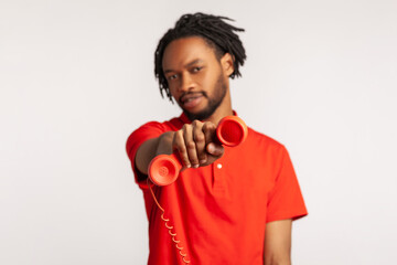 Good looking man with dreadlocks wearing red casual style T-shirt, holding and showing retro phone handset to camera, asking to answer phone. Indoor studio shot isolated on gray background.