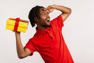 Side view of man with dreadlocks wearing red casual T-shirt, looking far away with hand over eyes, watching and waiting future birthday celebration. Indoor studio shot isolated on gray background.