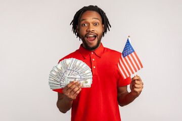 Amazed man wearing red casual style T-shirt, holding dollars banknotes and american fla, looking at camera with astonished surprised expression. Indoor studio shot isolated on gray background.