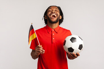 Screaming man wearing red casual T-shirt, football fan sincerely rejoicing, supporting favorite team holding ball and german flag, watching football. Indoor studio shot isolated on gray background. - obrazy, fototapety, plakaty
