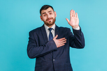 I promise to tell truth! Bearded man wearing official style suit standing raising hand and saying swear, making loyalty oath, pledging allegiance. Indoor studio shot isolated on blue background.