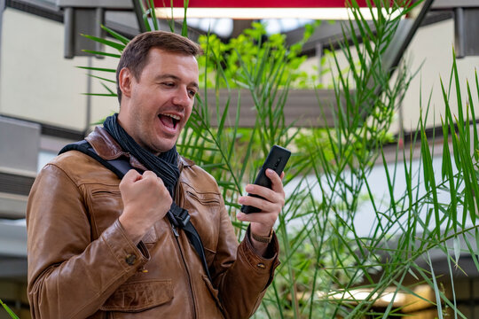Man with happy face in brown leather jacket check phone and stand in lobby against green plant at office recreational area. Excited man holding cellphone indoors. Happy man receive good news on phone