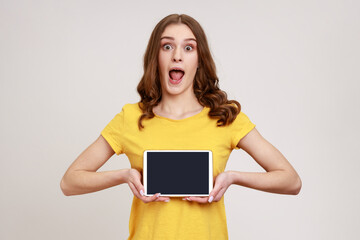 Portrait of shocked beautiful teenage girl in style T-shirt, showing tablet empty screen with unbelievable face, looking at camera with opened mouth. Indoor studio shot isolated on gray background.