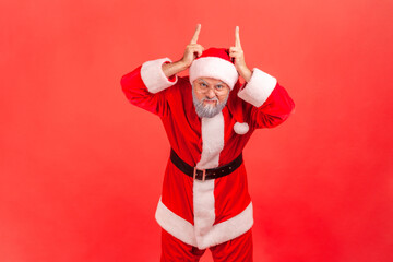 Portrait of elderly man with gray beard wearing santa claus costume showing bull horns on head, threatening to attack, danger and aggression. Indoor studio shot isolated on red background.