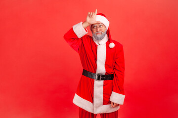 Fototapeta na wymiar Upset unlucky elderly man with gray beard wearing santa claus costume showing looser gesture holding finger near forehead, depressed with her fail. Indoor studio shot isolated on red background.