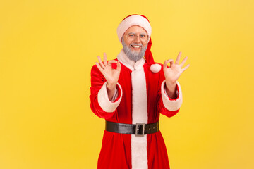 Fototapeta na wymiar Portrait of satisfied elderly man with gray beard wearing santa claus costume showing okay sign with both hands, looking at camera toothy smile. Indoor studio shot isolated on yellow background.