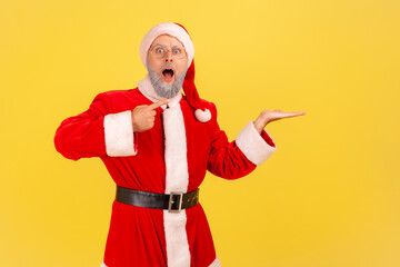 Fototapeta na wymiar Shocked elderly man with gray beard wearing santa claus costume pointing at his palm, presenting mock up, showing astonished advertisement. Indoor studio shot isolated on yellow background.