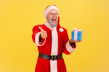 Excited positive elderly man with gray beard wearing santa claus costume with blue wrapped present box. showing thumb up to camera with toothy smile. Indoor studio shot isolated on yellow background.