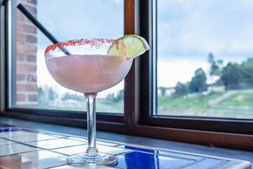 Alcoholic cocktail Margarita with clear Blanco tequila and lemon in a glass, the edges of which are sprinkled with salt on the windowsill by the window