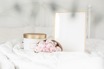 Fototapeta na wymiar Gold gift box and white frame mockup on the bed. Bouquet of white peonies in craft packaging. Scandivanavian white interior.