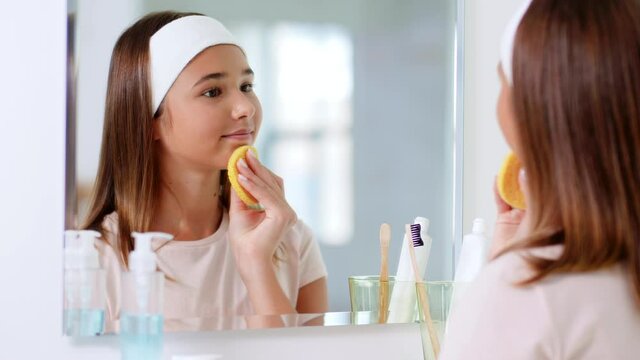 beauty, hygiene and people concept - teenage girl with cleansing sponge cleaning facial skin and looking in mirror at bathroom