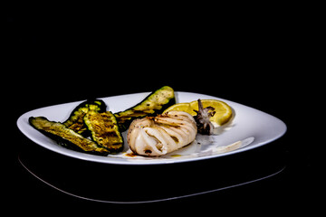 mediterranean dish with roasted squid and zucchini and lemon, on black reflective background