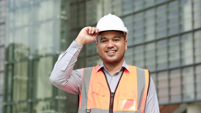 Smiling Asian engineer handsome man or architect looking camera with white safety helmet in construction site. Standing at modern building construction. Worker asian man working project building