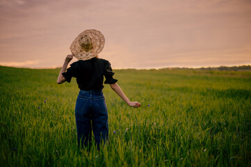Rear, back view of fashionable woman wearing straw hat, black vintage shirt, blue denim jeans, posing in summer green field. Copy, empty space for text