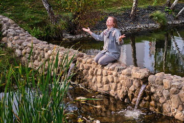 A happy middle-aged woman smiles sitting on the rocks by the river. An adult woman enjoys nature sitting in the rays of the summer sun