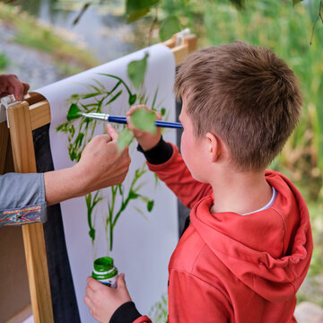 Mother teaches boy son to paint on canvas. Woman teacher artist paints with a child on paper nature and trees by the river