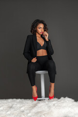 Portrait of a sensual black woman with curly dark hair and beautiful makeup sitting by herself on a bed of fur inside a studio with a grey background wearing a black jacket with red high heels.