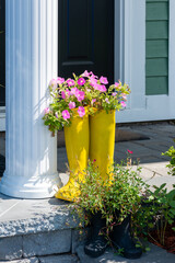 The entrance of a house with a black door, green exterior wall, white pillar. On the step are two yellow rubber boots with pink flowers and two children's rubber boots with green foliage growing out. 