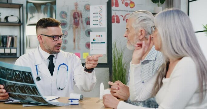 Attractive positive skilled bearded doctor explaining results of x-ray scan to confident senior couple which visiting him in medical office