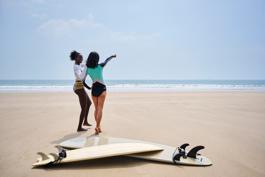 Smiling multiracial girlfriends pointing away against surfboards on sea shore