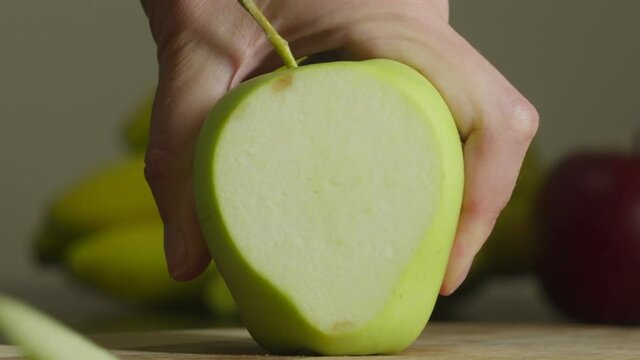 Close-up of hand holding appetizing green apple, cutting slice of fruit with kitchen knife on blurred background of tropic fruits indoors. Cut piece of fruitage falling off on chopping board