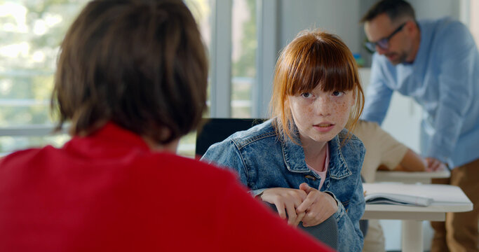 Rear View Of Teen Girl Student Turn Back And Talk To Classmate Sitting At Desk On Classroom