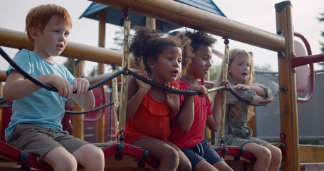 Multiethnic preschool children sit on suspension bridge on playground - obrazy, fototapety, plakaty