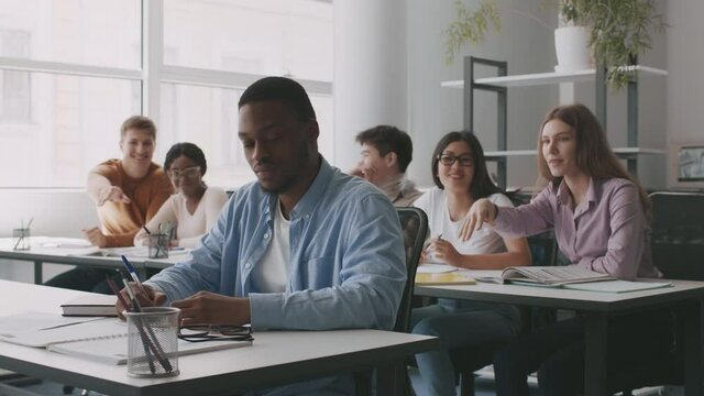 Young african american guy suffering from groupmates, sitting apart in classroom, students throwing paper balls at him