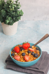 A blue bowl of refreshing mixed berries cereal and a metal spoon with a plant in a white vase.