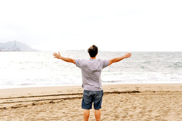 young adult man on the beach looking at the sea from behind with open arms with feeling of freedom