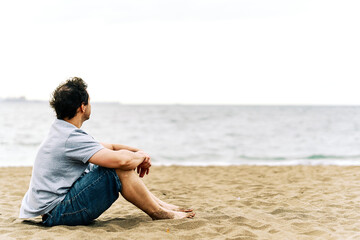Fototapeta na wymiar Thoughtful young adult man sitting on the beach sand with melancholic gesture