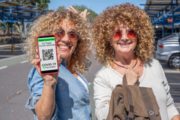 Two smiling curly women ready to travel holding mobile phone with digital certificate of vaccination of covid 19