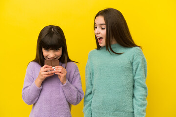 Little sisters isolated on yellow background eating a chocolate tablet