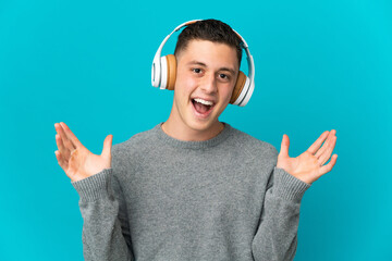 Young caucasian man isolated on blue background surprised and listening music