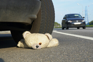 Children's toy - a teddy bear lies on the road under the wheel of a car against the background of another passing car. The concept of road accidents and road hazards.