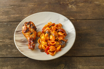 Seashell pasta in tomato with chicken leg baked on the grill on a wooden table. Horizontal photo