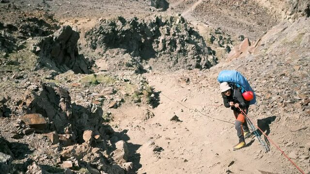 A climber with a large backpack descends a steep slope in the mountains holding on to a rope. A tourist overcomes a difficult section holding on to a rope. Mountain climber