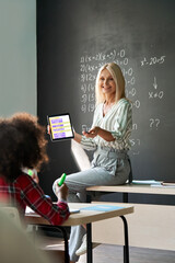 Smiling female teacher demonstrating new maths task holding using tablet device to elementary...
