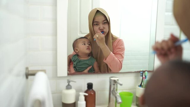 muslim mother brushing her teeth while holding her baby son