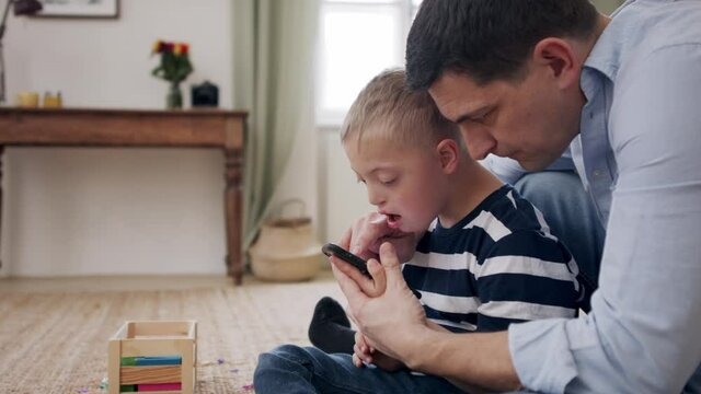 Father with happy down syndrome son indoors at home, using smartphone.