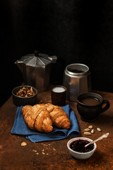 French breakfast. Freshly baked croissants, a cup of coffee, jam and nuts on an aged rusty background. Dark style still life concept with copy space. 