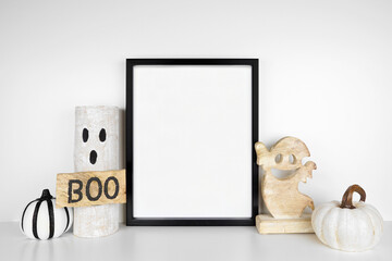 Halloween mock up. Black frame on a white shelf with rustic wood ghost decor and pumpkins. Portrait frame against a white wall. Copy space.
