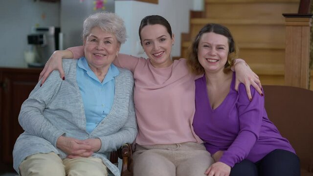 Portrait of three positive Caucasian women of different ages sitting on couch at home looking at camera smiling. Happy grandmother mother and daughter posing indoors. Generations and family