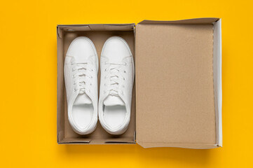 White women's leather sneakers in cardboard box on yellow background top view flat lay. Stylish youth sneakers, sports shoes, genuine leather footwear. Minimalistic shoe store advertising, fashion