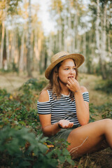 Beautiful young girl sitting and eating blackberries in the woods. Girl in a straw hat in the woods