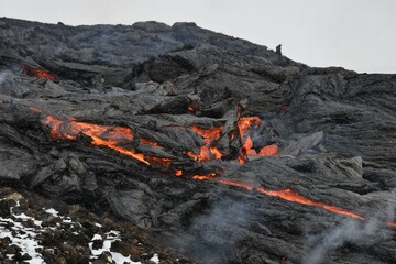 A slab of lava crust is overturned in a lava flow at Fagradalsfjall, Iceland. The lava surface is black, with red, molten lava visible beneath and steam rising. Snowy hills in the background.